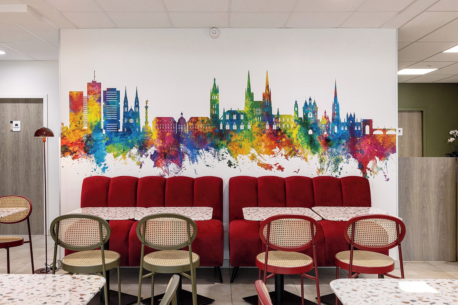 A restaurant with red upholstered booths, wicker chairs, and a colorful cityscape mural on the wall.