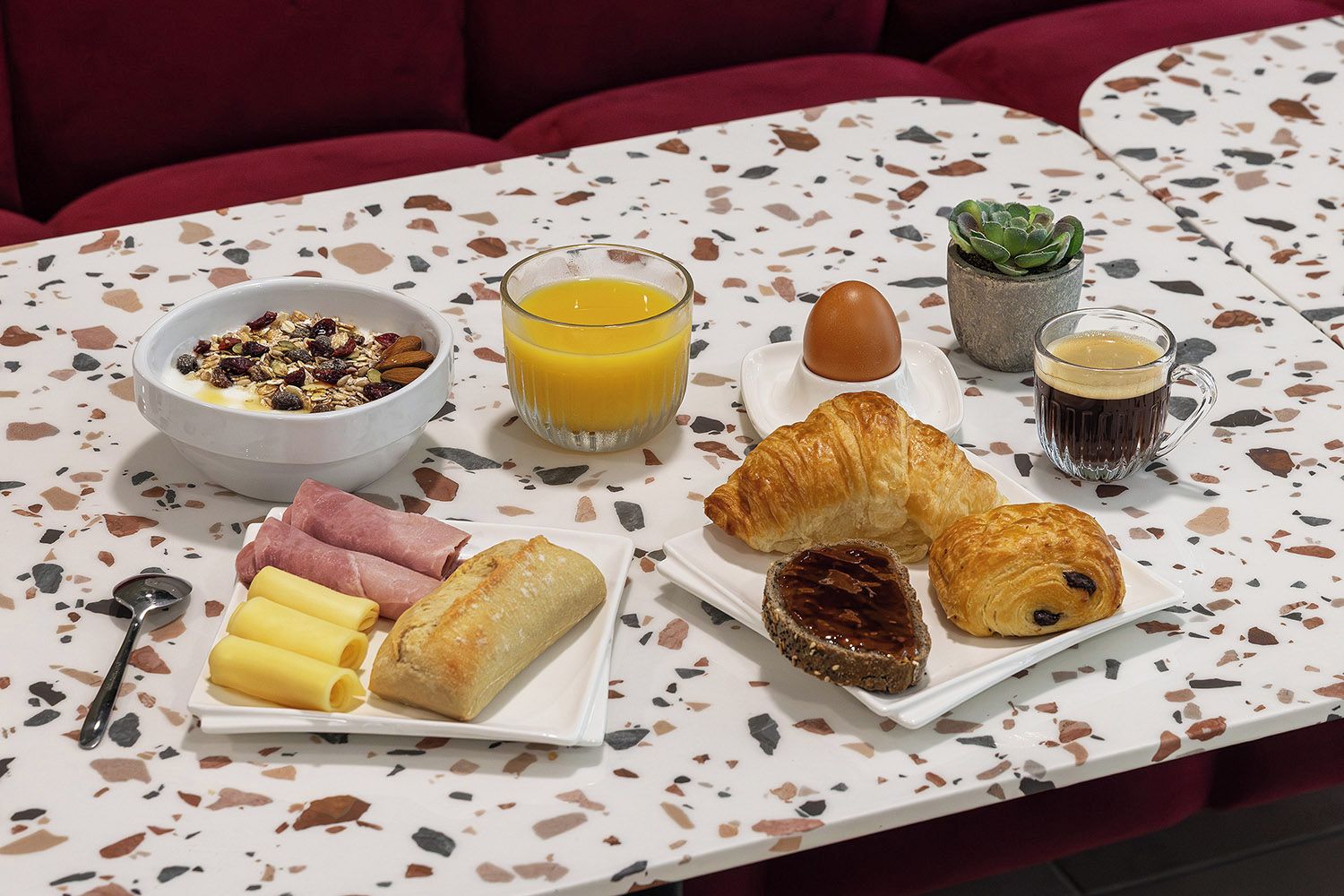 A breakfast table with croissants, a bowl of cereal, a glass of orange juice, a cup of coffee, and a small potted plant.