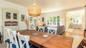 A dining area with a wooden table, white chairs, a hanging light fixture, and a kitchen area with cabinets and a window. - Cottage with forest idyll and peace by Arresø (Frederiksværk)