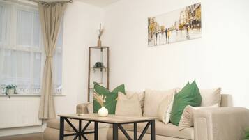 A living room with a beige sofa, a wooden coffee table, a tall plant stand, and a cityscape painting on the wall.