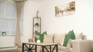 A living room with a beige sofa, a wooden coffee table, a tall plant stand, and a cityscape painting on the wall. - Luxury 3BR Home. 20 Mins to London. Garden + Wi-Fi. Sleeps 8 (Gravesend)
