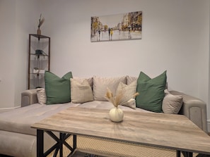 A living room with a sofa, a coffee table, a painting on the wall, and a sideboard with plants. - Luxury 3BR Home. 20 Mins to London. Garden + Wi-Fi. Sleeps 8 (Gravesend)