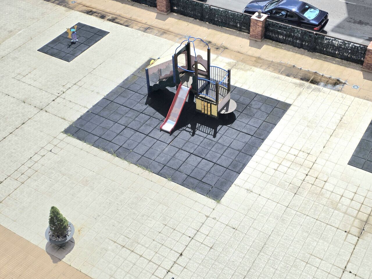 Un’area giochi con scivolo e struttura per arrampicata, circondata da una superficie pavimentata e un vaso con una pianta.