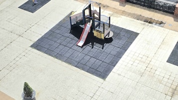 Un’area giochi con scivolo e struttura per arrampicata, circondata da una superficie pavimentata e un vaso con una pianta.