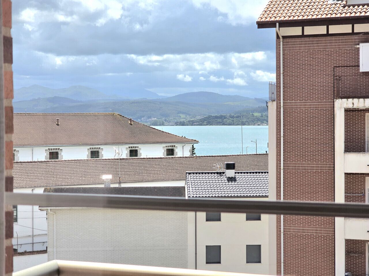 Vista di una cittadina costiera con edifici dalle facciate in terracotta, una distesa d'acqua e montagne in lontananza.