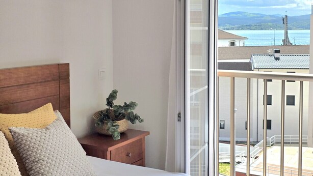 Une chambre à coucher avec un lit, une table de chevet, une plante en pot et une vue sur un balcon doté d’une balustrade, avec un immeuble en arrière-plan.