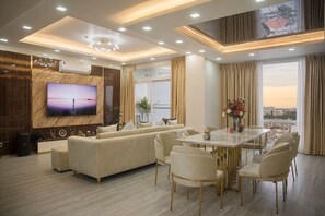 A modern living room with a large TV, a sofa, a dining table with chairs, and a city view. - The Crown Luxury 4BR Penthouse (Phu Nhuan)
