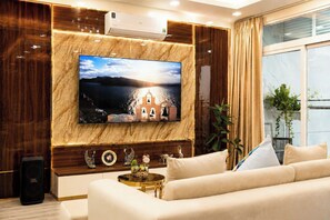 A modern living room with a large flat-screen TV mounted on a marble wall, a sofa, a coffee table, and a speaker. - The Crown Luxury 4BR Penthouse (Phu Nhuan)