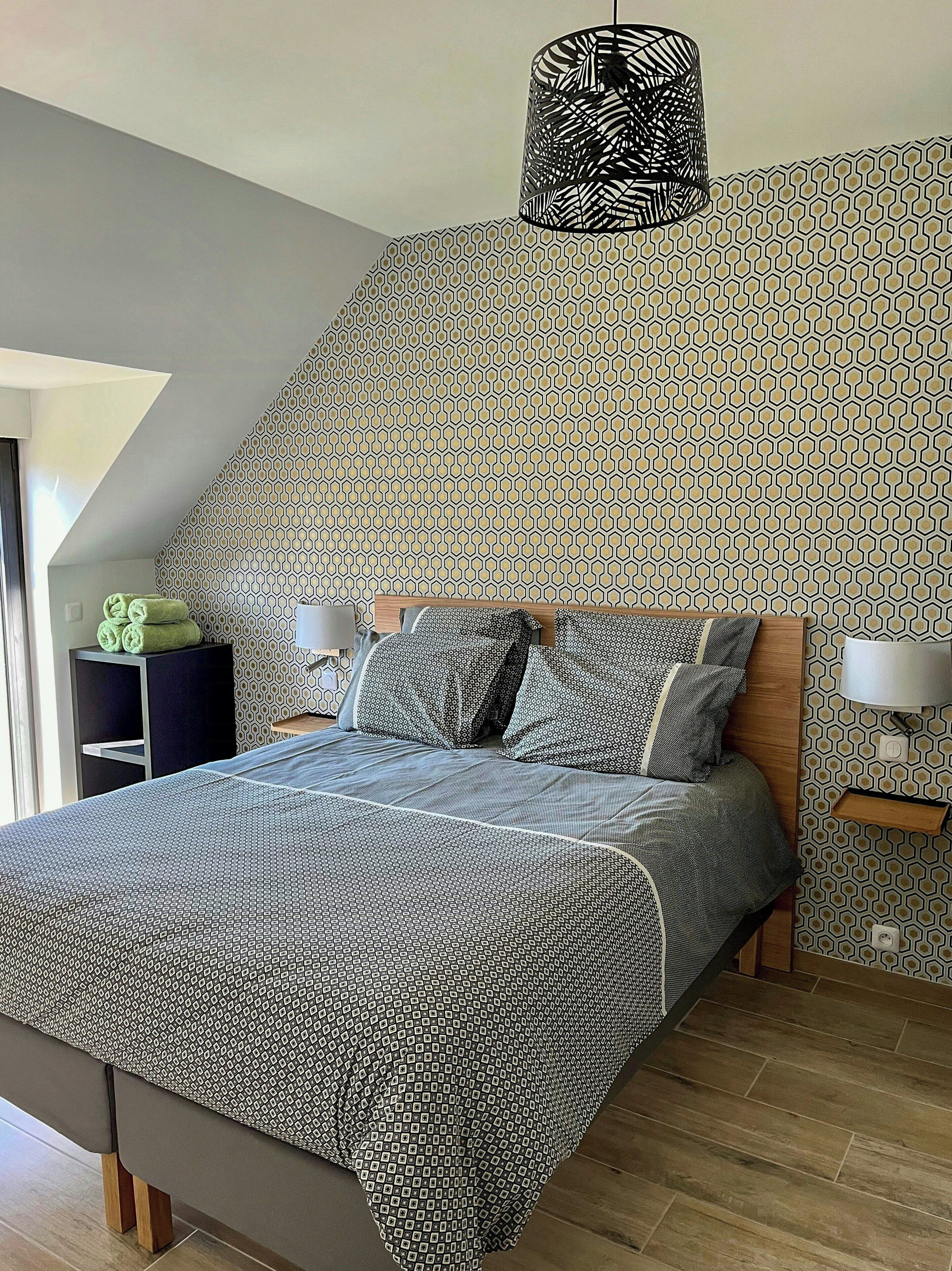 Een slaapkamer met een behang met patroon, een bed met een bedovertrek met patroon, een bedlamp en een plafondlamp.