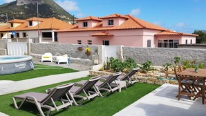Ein Garten mit Swimmingpool, Liegestühlen, Grillplatz und Blick auf rosa Häuser.