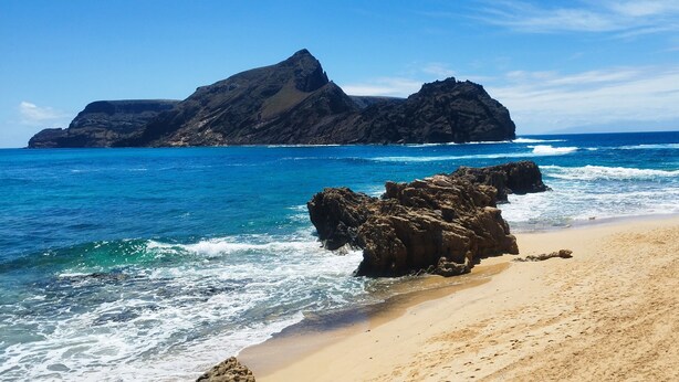 Ein Strand mit klarem, blauem Wasser, Felsformationen und einem Sandufer.