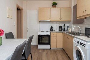A compact kitchen with a stove, oven, microwave, and washing machine, wooden cabinets, and a dining area with chairs. - Villy Suites II - Zante Lux Suite Near the Beach (Argassi)