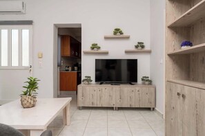 Ein modernes Wohnzimmer mit Flachbildfernseher auf einem Holzschrank, einem Esstisch mit Pflanze und einer durch einen offenen Durchgang einsehbaren Küche.