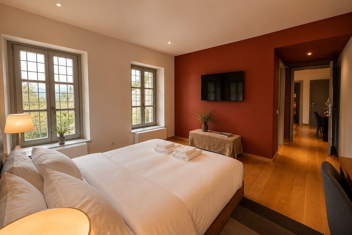 Un dormitorio con una cama grande, dos ventanas, una mesita de noche con una planta, un televisor en la pared y un pasillo que conduce a otra habitación.