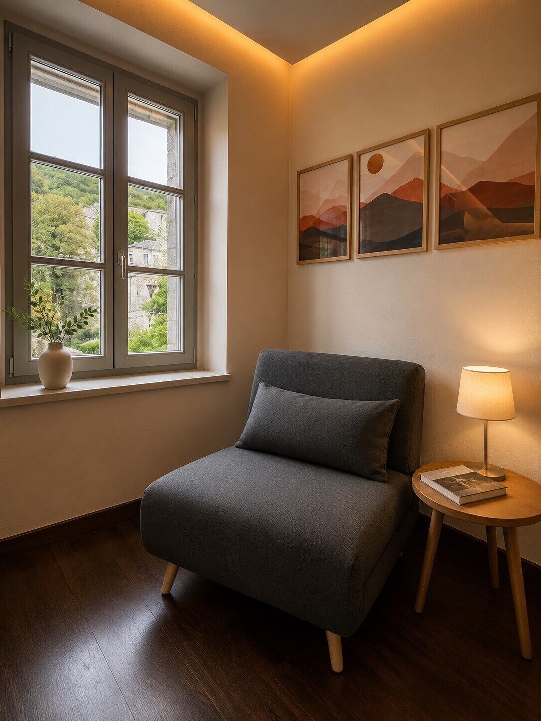 A room with a grey sofa, a small round table with a lamp, a window with a view of greenery, and three framed pictures on the wall.