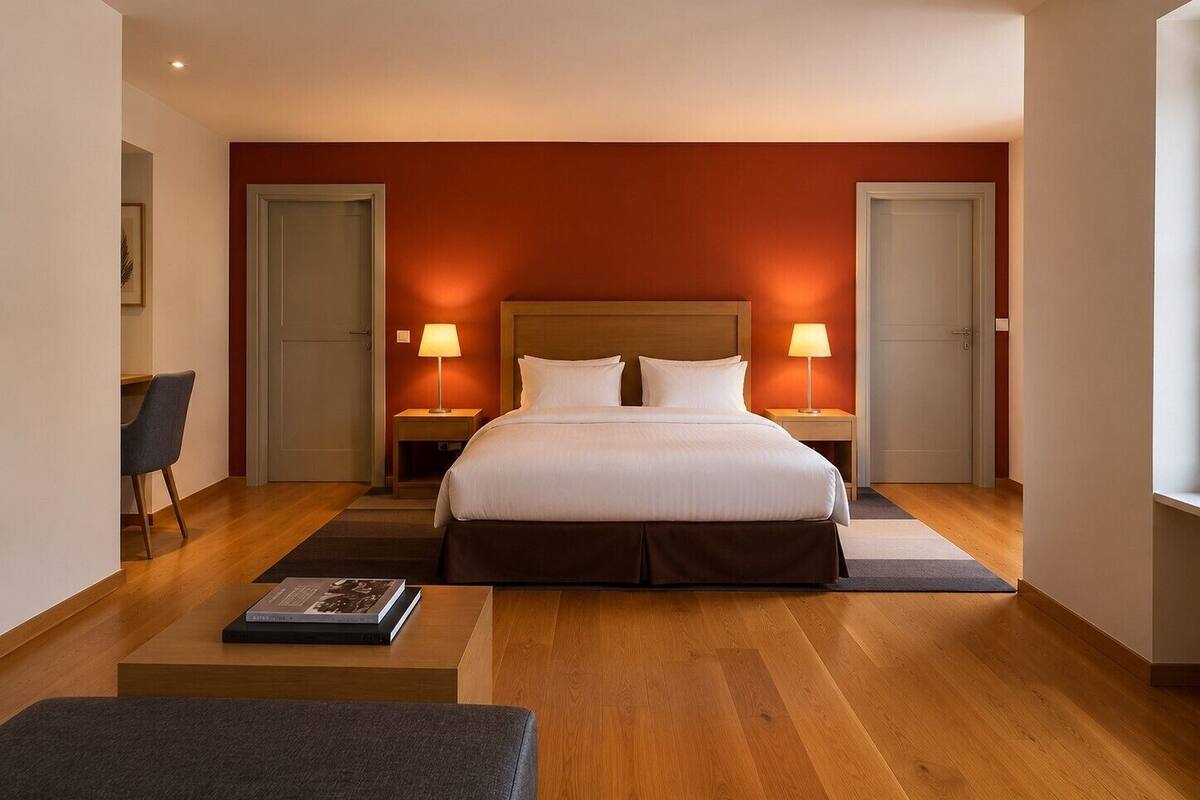 Una habitación de hotel con una cama grande, dos mesitas de noche con lámparas, una silla, una pequeña mesa con un libro y suelo de madera.