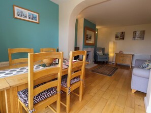 A dining area with wooden chairs and a table, a living room with a sofa and armchair, and a kitchen area with cabinets and a countertop. - Little Gem (Weymouth)