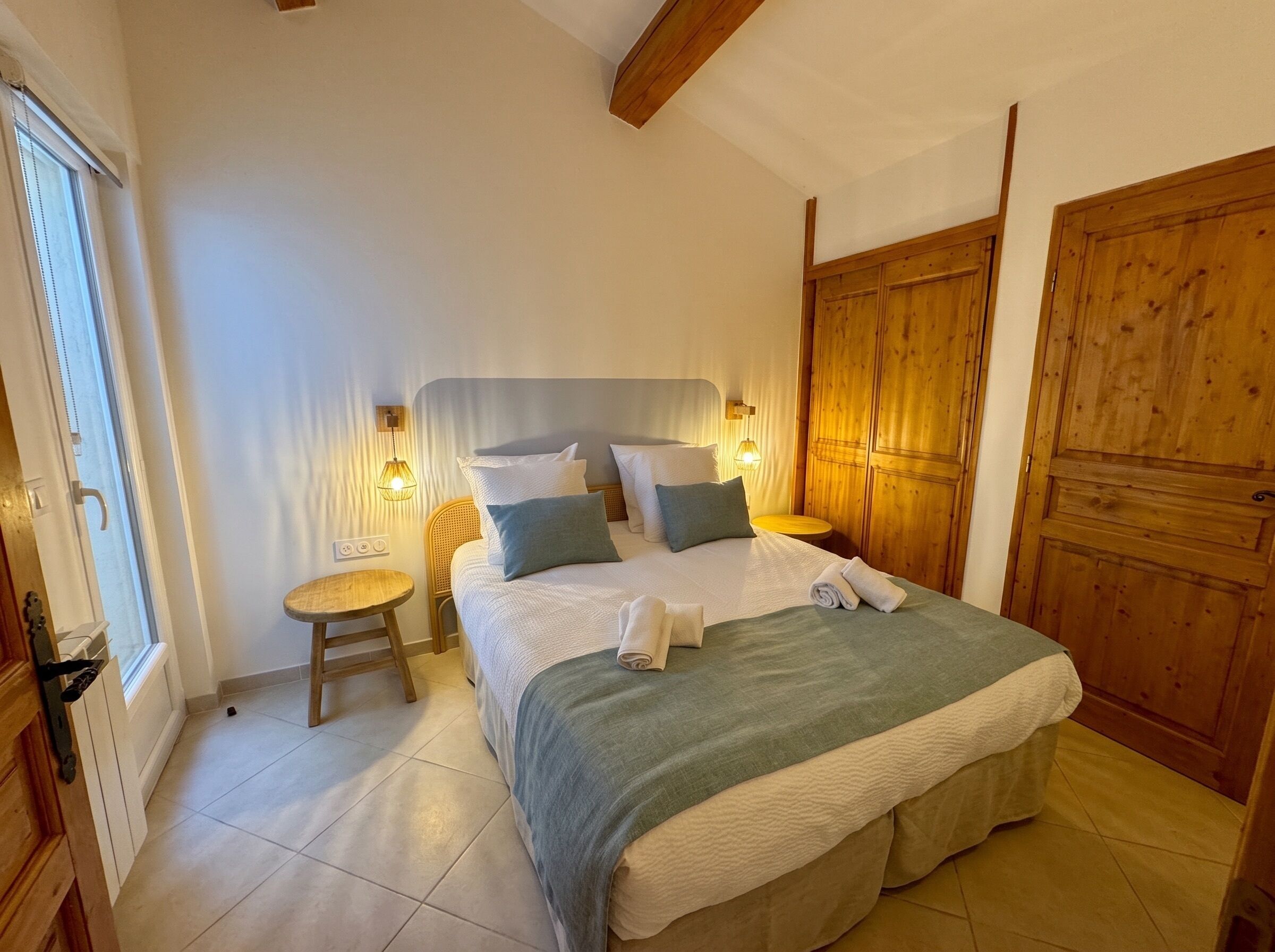 A bedroom with a large bed, two bedside tables, a wooden wardrobe, and a window with blinds.