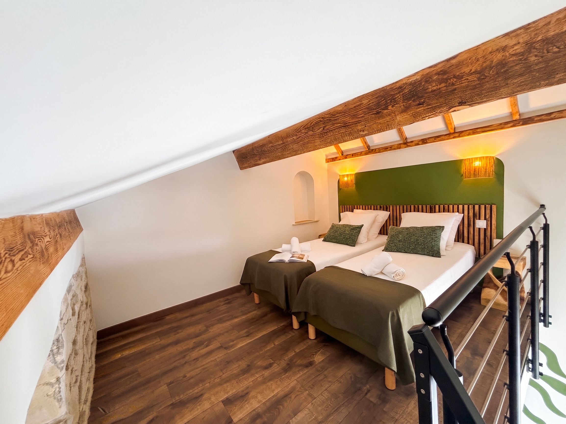 A room with two beds, wooden flooring, a green accent wall, and a staircase.