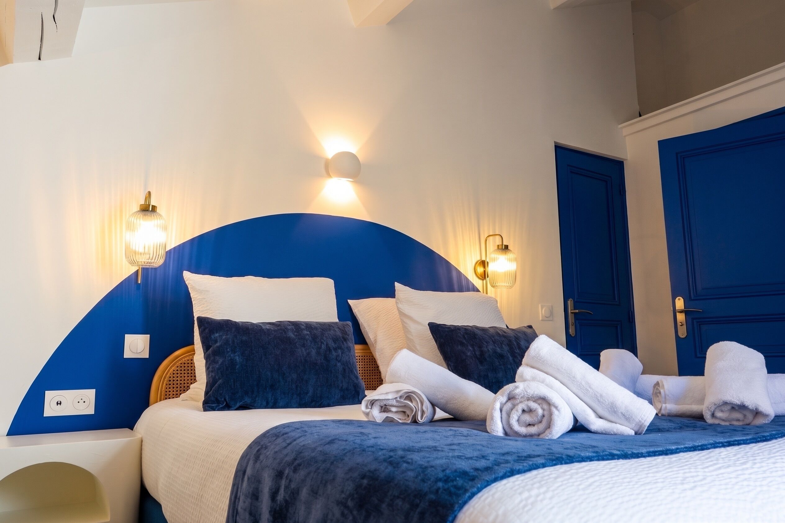 A hotel room with a bed, two wall-mounted lights, a blue door, and a power outlet on the wall.