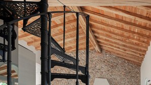 Un escalier en colimaçon, une cheminée en pierre et un canapé d’angle dans une pièce aux poutres apparentes et au mur en pierre.