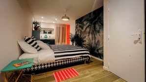 Een slaapkamer met een bed, een tafeltje, een rood kleed, een deur en een muur met een palmboom muurdecoratie.