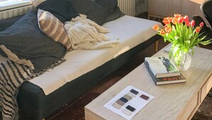 Een knus ingerichte kamer met een bed, een houten bijzettafeltje met boeken en een vaas, en uitzicht op groen.