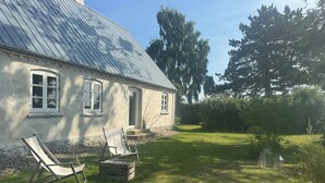 Een huis met een schuin metalen dak, een voordeur en een klein portaal. Op het gras staan twee tuin stoelen en een houten kist.