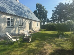 Ein Haus mit einem geneigten Metalldach, einer Haustür und einer kleinen Veranda. Auf dem Rasen stehen zwei Gartenstühle und eine Holzkiste.