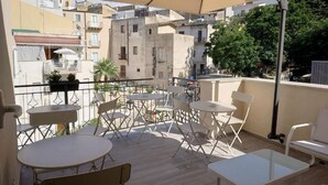 A balcony with white foldable chairs and tables, a beige umbrella, and a view of buildings and trees. - MARINA BLU ROOMS (Castellammare del Golfo)