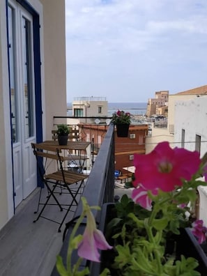 Ein Balkon mit Tisch und Stühlen, Blumentöpfen und Blick auf das Meer und Gebäude.