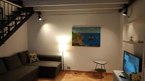 A living room with a sofa, a small table, a TV, a painting, and a staircase. - MARINA BLU ROOMS (Castellammare del Golfo)
