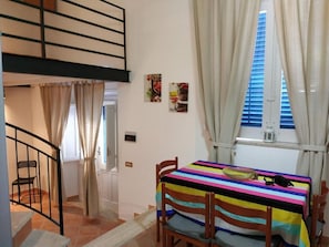 A dining area with a table covered by a striped tablecloth, wooden chairs, a window with curtains, and a staircase leading to an upper level.