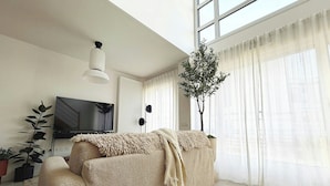 Ein Wohnzimmer mit einer Couch, einem Fernseher, einer Pflanze und einem Fenster mit Vorhängen.