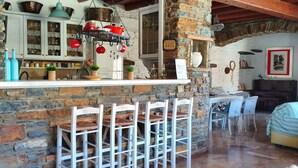 A rustic kitchen with stone walls, wooden beams, and a bar area with white chairs. - Sun Holiday Kea Private Pool Sea View (Kea)
