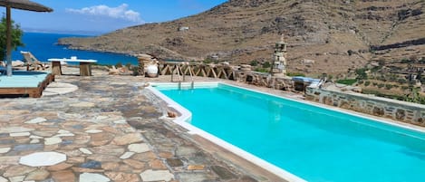 Piscina com piso de pedra, espreguiçadeiras e montanhas ao fundo.