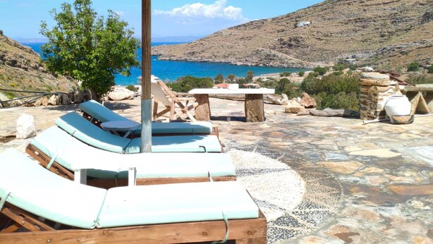 A stone patio with lounge chairs and a table, overlooking a body of water and hills. - Sun Holiday Kea Private Pool Sea View (Kea)