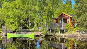 A cabin with a wooden deck, a canoe, and a dock extending into a calm lake surrounded by trees. - Lakeside cabin with paddle board (Lerum)