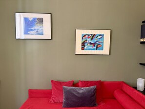 Ein Zimmer mit einem roten Sofa, zwei gerahmten Kunstwerken und einer weißen Vase auf einem Beistelltisch.