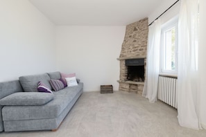 Ein Wohnzimmer mit Kamin aus Stein, einem grauen Sofa mit Kissen und einem Fenster mit weißen Vorhängen.