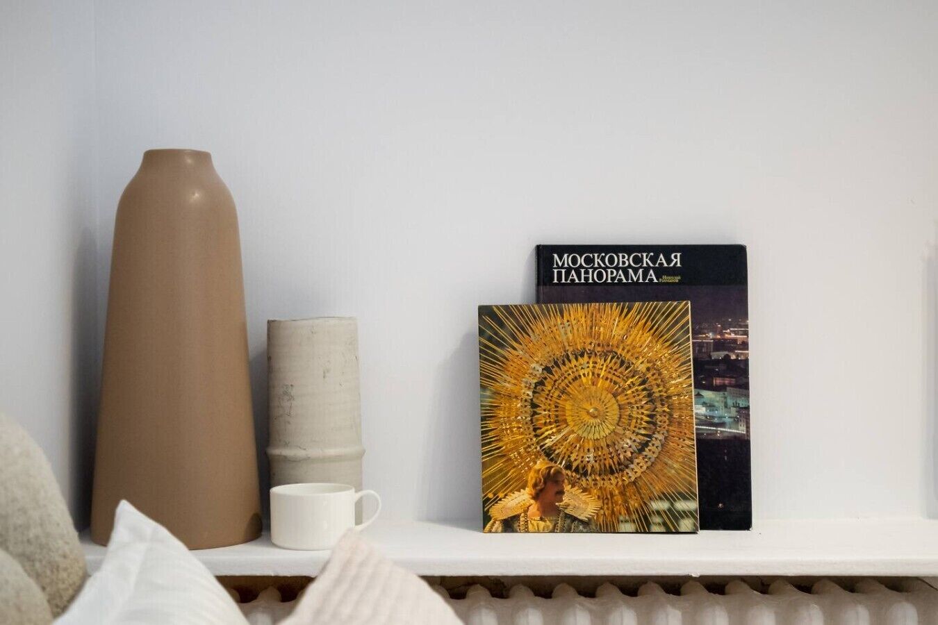 Uno scaffale bianco con un vaso alto, un contenitore cilindrico, una tazza bianca e un libro intitolato 'МОСКОВСКАЯ ПАНОРАМА'.