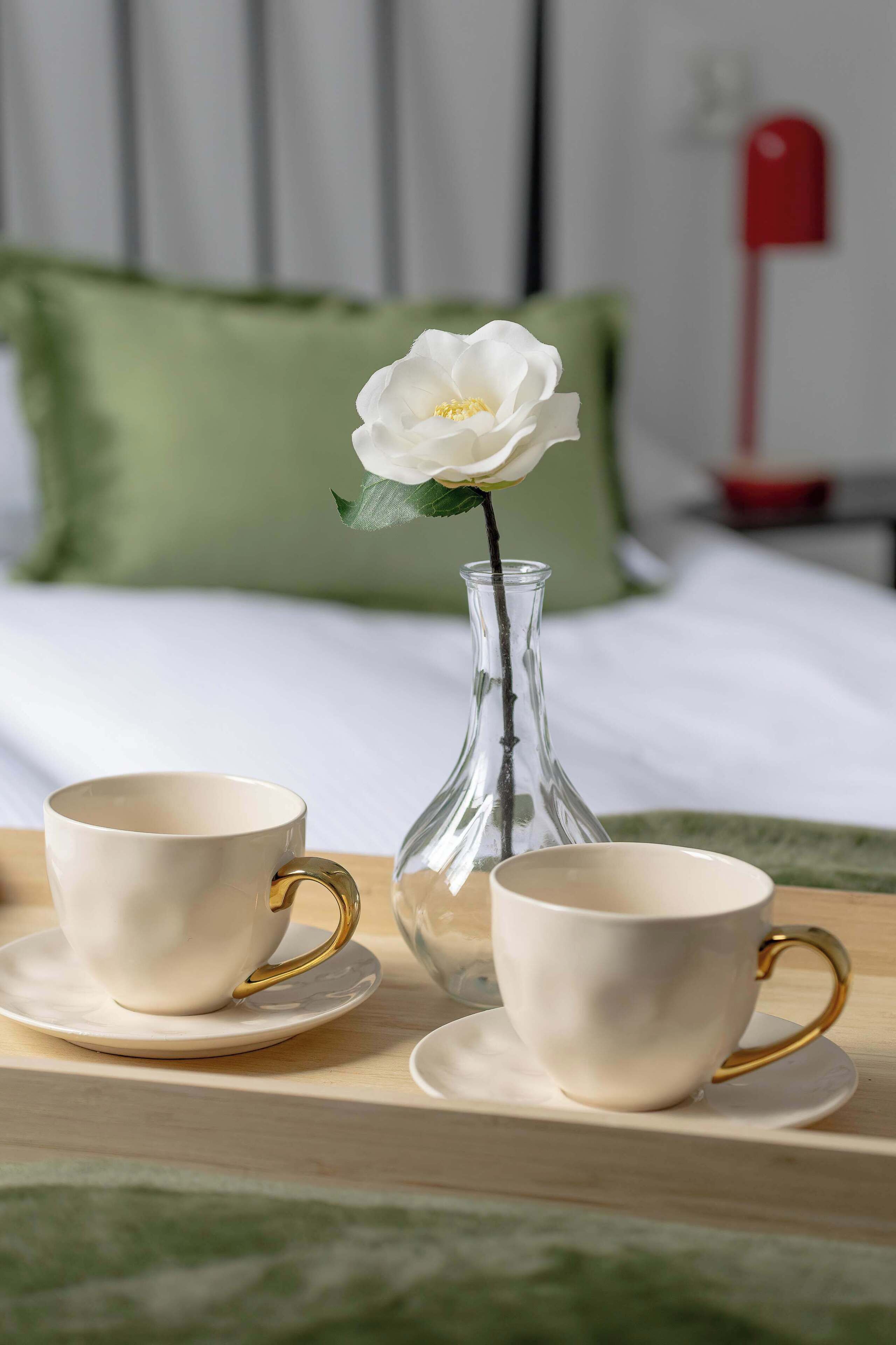 A bed with a green pillow, a white vase with a flower, and two white cups with gold handles on a tray.