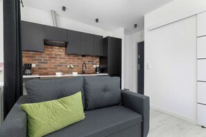 Ein moderner Wohnbereich mit grauem Sofa, einem grünen Kissen, einer Wandverkleidung mit Ziegeloptik und einer schwarzen Küche mit dunklen Schränken.