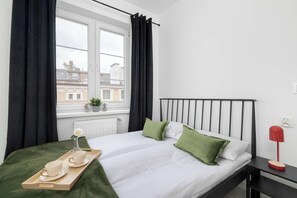 Ein Schlafzimmer mit Bett, Nachttisch, einem Fenster mit Vorhängen und einem Tablett mit Tassen und einer Blume.