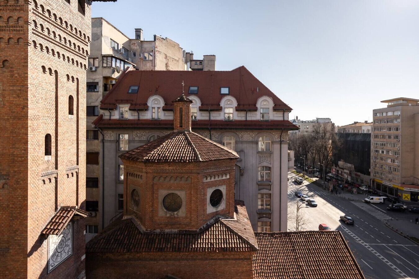 Un bâtiment historique doté d’un clocher, d’un toit recouvert de tuiles rouges et donnant sur une rue animée où l’on peut voir des voitures et d’autres bâtiments.