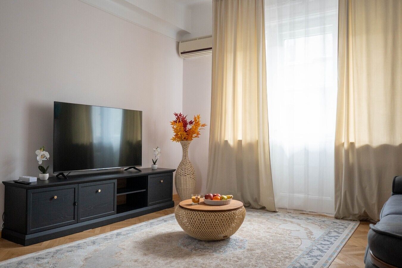 Un salon moderne avec un téléviseur à écran plat posé sur un meuble en bois foncé, une table basse ronde en fibres naturelles sur laquelle sont disposés des fruits, un vase contenant des fleurs séchées et une grande fenêtre ornée de rideaux légers.
