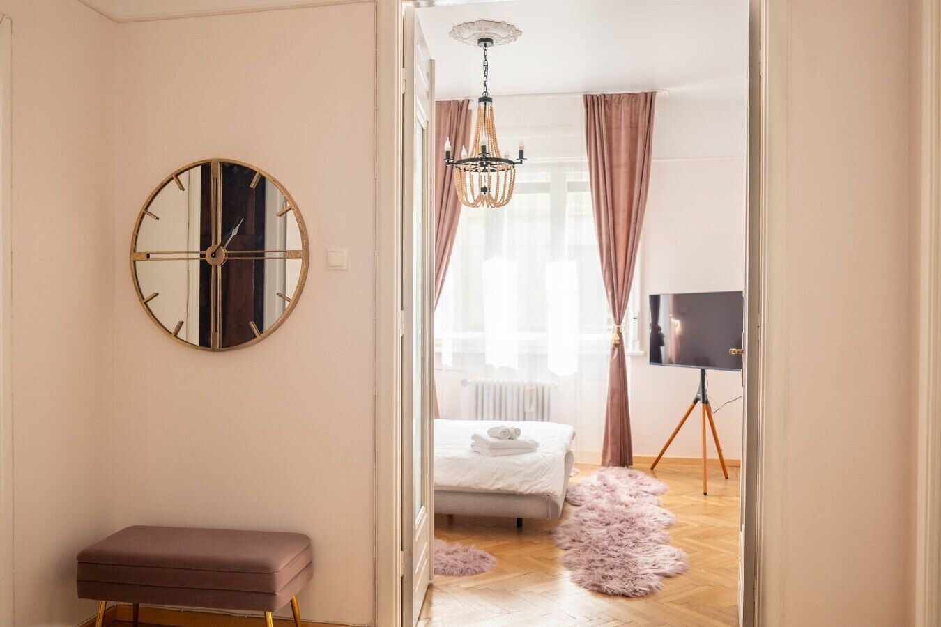 A room with a round mirror, a bench, a bed, a television on a tripod, and a chandelier.