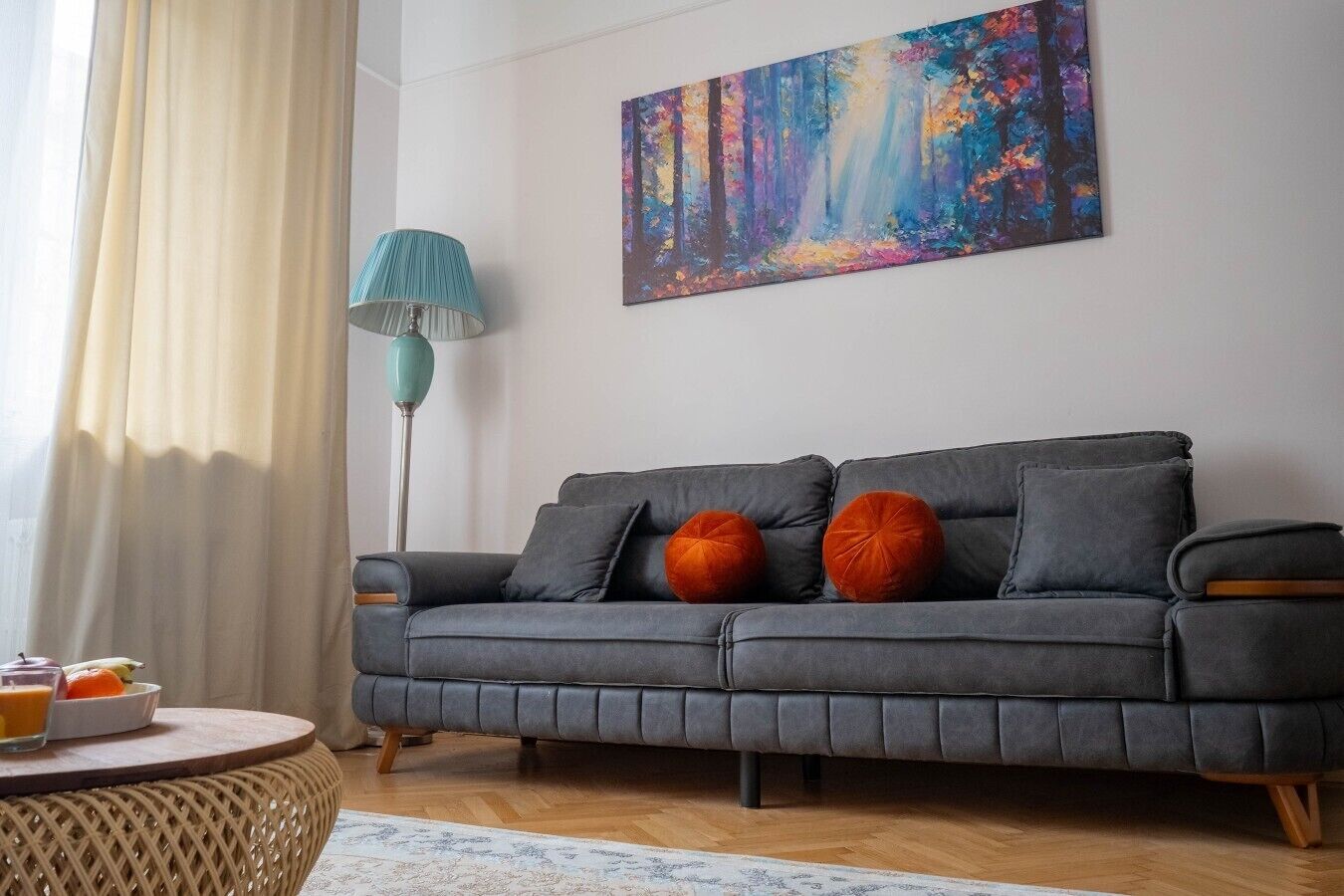 A modern living room with a grey sofa, a colorful abstract painting, a teal floor lamp, and a round coffee table with a bowl of fruit.