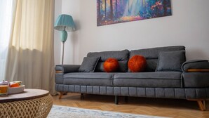 A modern living room with a grey sofa, a colorful abstract painting, a teal floor lamp, and a round coffee table with a bowl of fruit. - Luxury l Apartment | Radisson | Calea Victoriei (Bucharest)