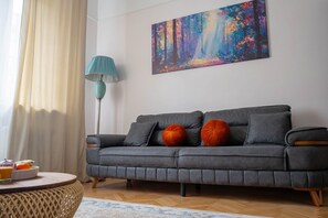 Ein modernes Wohnzimmer mit einem grauen Sofa, einem farbenfrohen abstrakten Gemälde, einer petrolfarbenen Stehlampe und einem runden Couchtisch mit Obstschale.